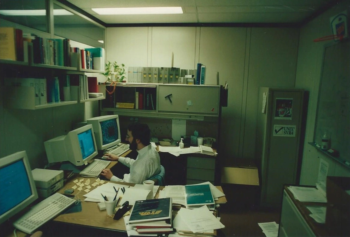 A programmer works at one of three CRT monitors in a cluttered 1994 office at Unix System Laboratories, surrounded by manuals, sticky notes, and a "UNIX SVR4" sticker on a cabinet